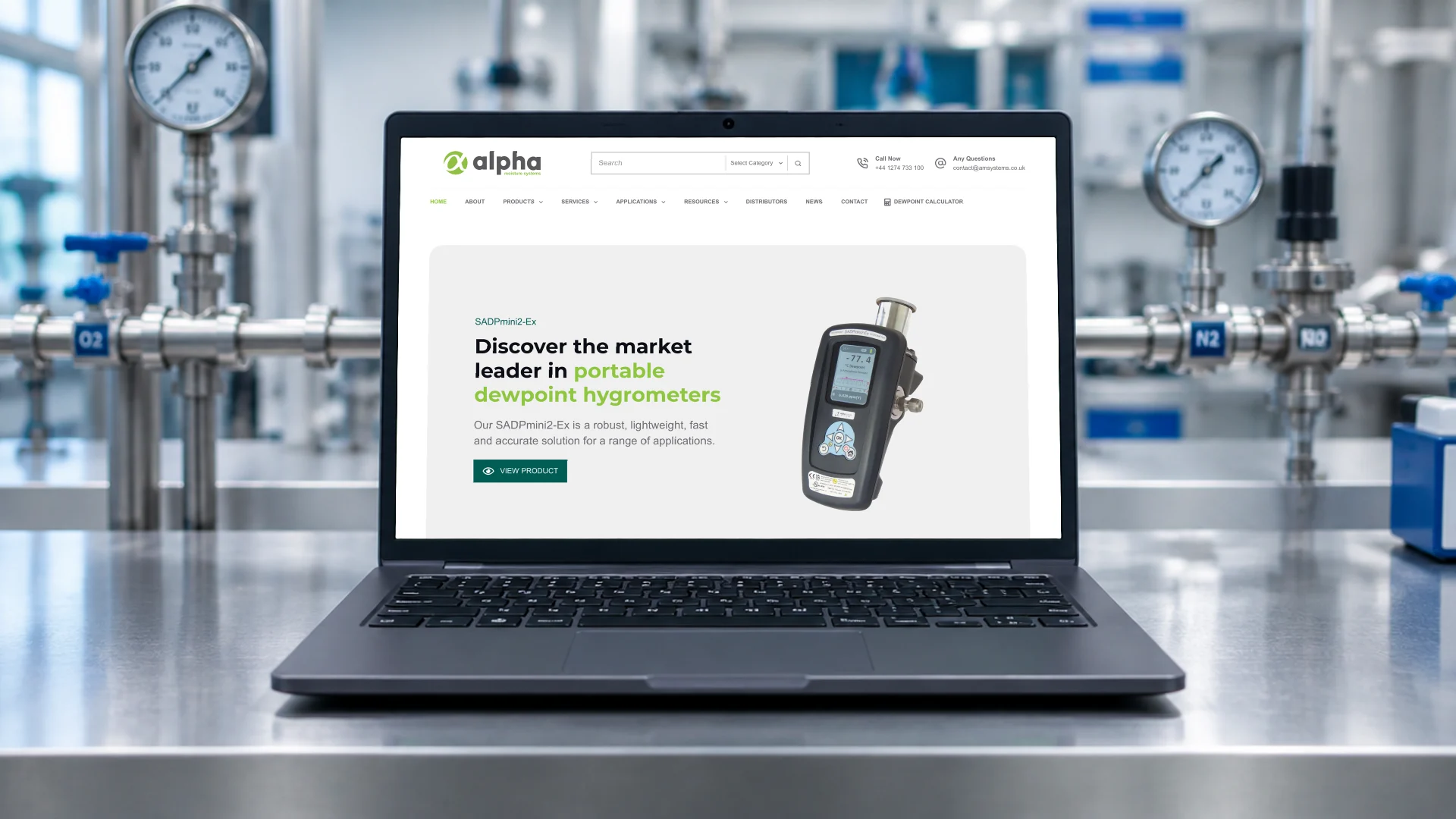 A laptop on a laboratory bench beside gas pipework and gauges, showing the Alpha Moisture Systems homepage and its portable dewpoint hygrometer