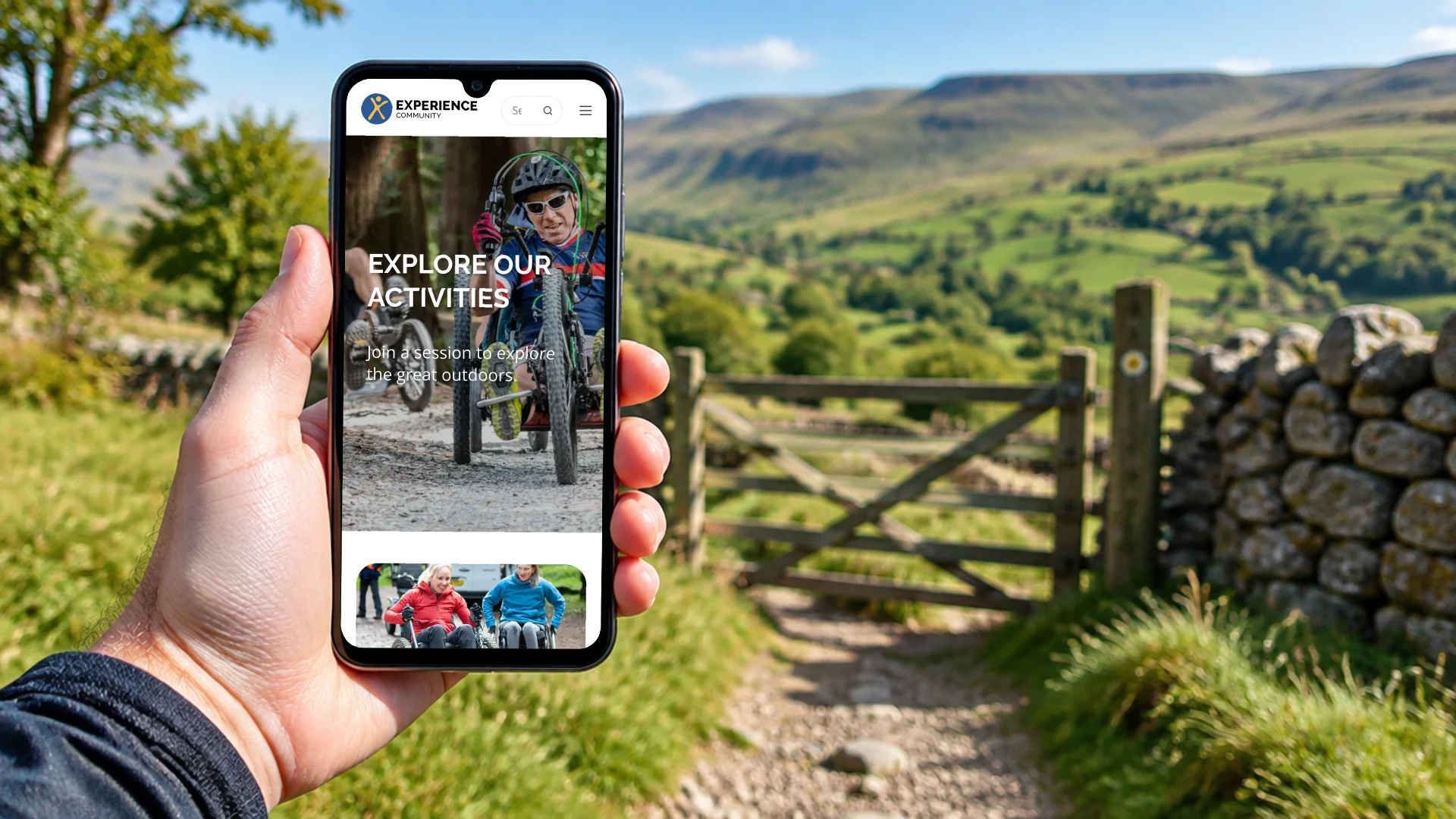 A hand holding a phone on a countryside trail by a dry stone wall, showing the Experience Community activities page on mobile