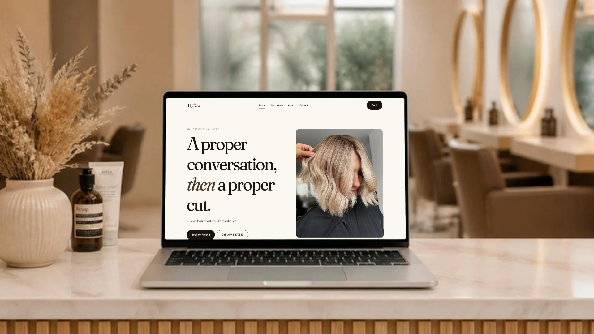 A laptop on a salon reception desk showing the H&Co Hairdressing homepage: serif headline, warm cream palette and the salon's own client photography