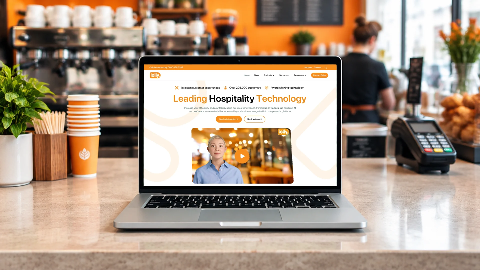 A laptop on a coffee shop counter beside a card terminal, showing the Lolly homepage: Leading Hospitality Technology