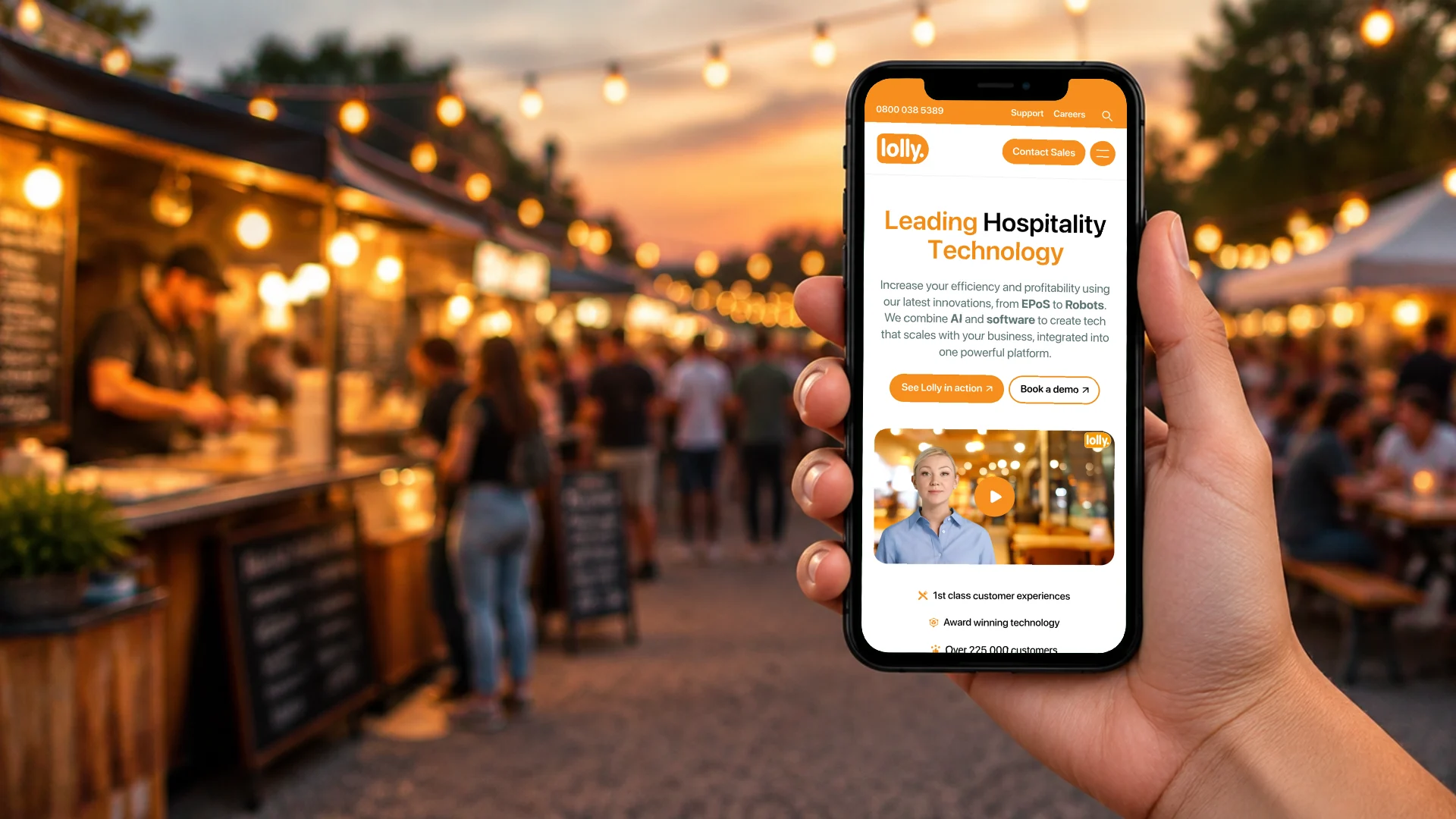 A hand holding a phone at an evening food festival, showing the Lolly homepage on mobile