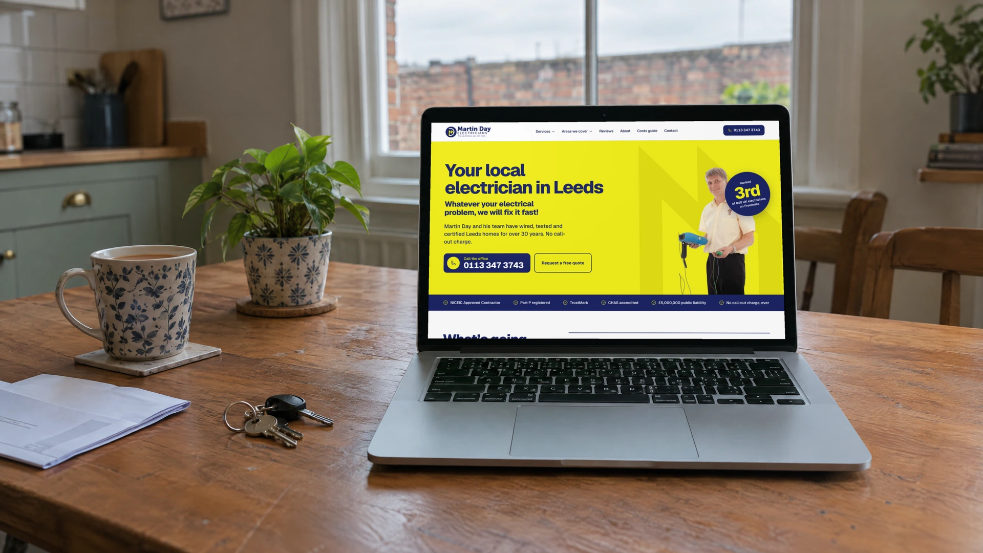 A laptop on a kitchen table showing the Martin Day Electricians homepage: yellow hero, Martin's own photo, the FreeIndex ranking badge and a bar of trade accreditations