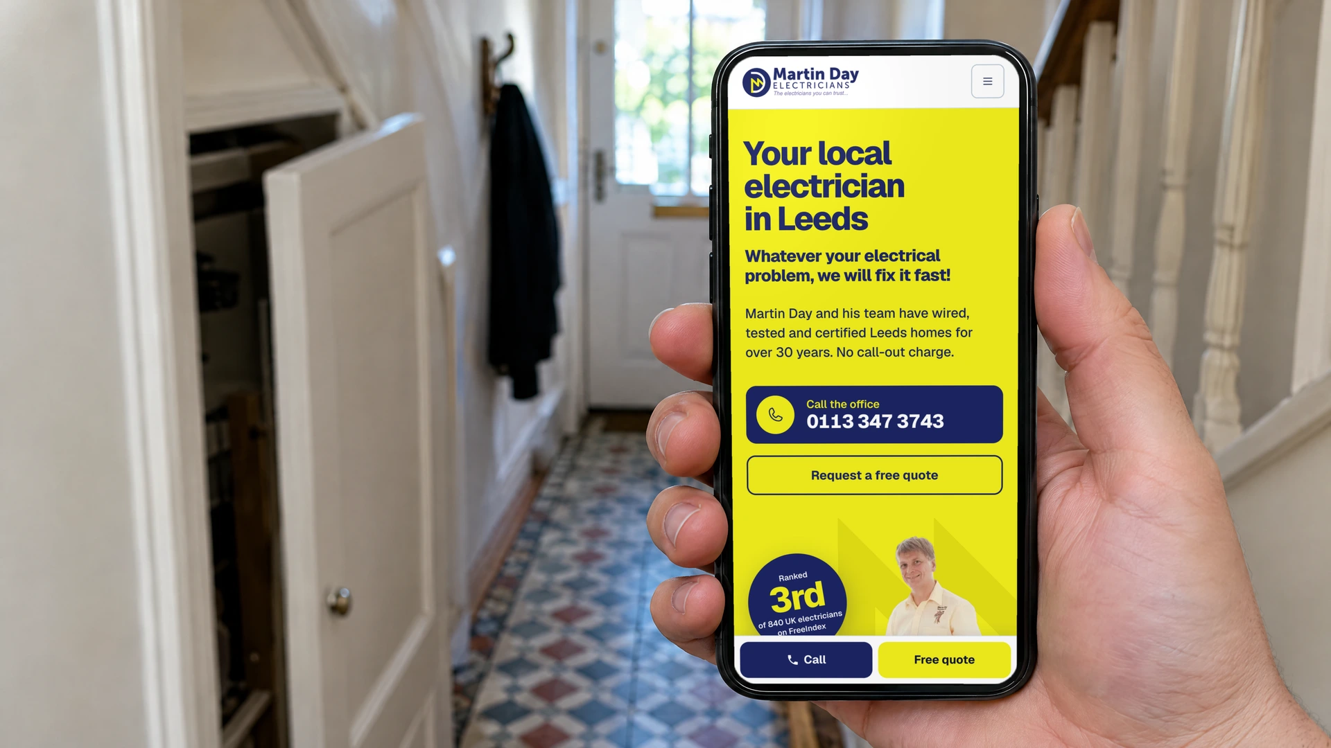A hand holding a phone in a terraced house hallway, showing the Martin Day Electricians homepage with the office number, free quote button and sticky call bar